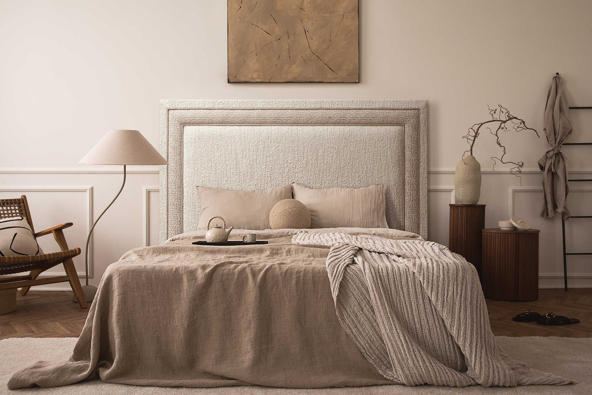 Ambiente de dormitorio elegante de estilo Boho. Aparece una cama, un cabecero tapizado en tela Boucle, una lámpara, una descalzadora, unas mesitas de noche, un jarrón y un cuadro en la pared.